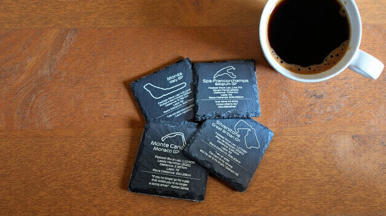 Four black stone coasters with white text on a wooden surface next to a cup of coffee.